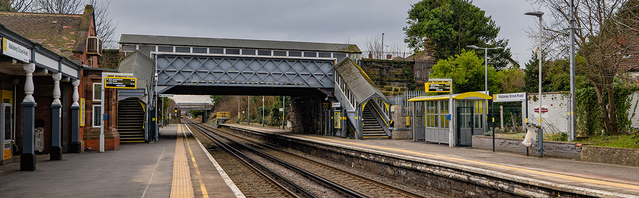 The platform at Wallasey Grove Road station. A pedestrian footbridge appears above the railway track. Sheltered seating areas also appear on the platform. 