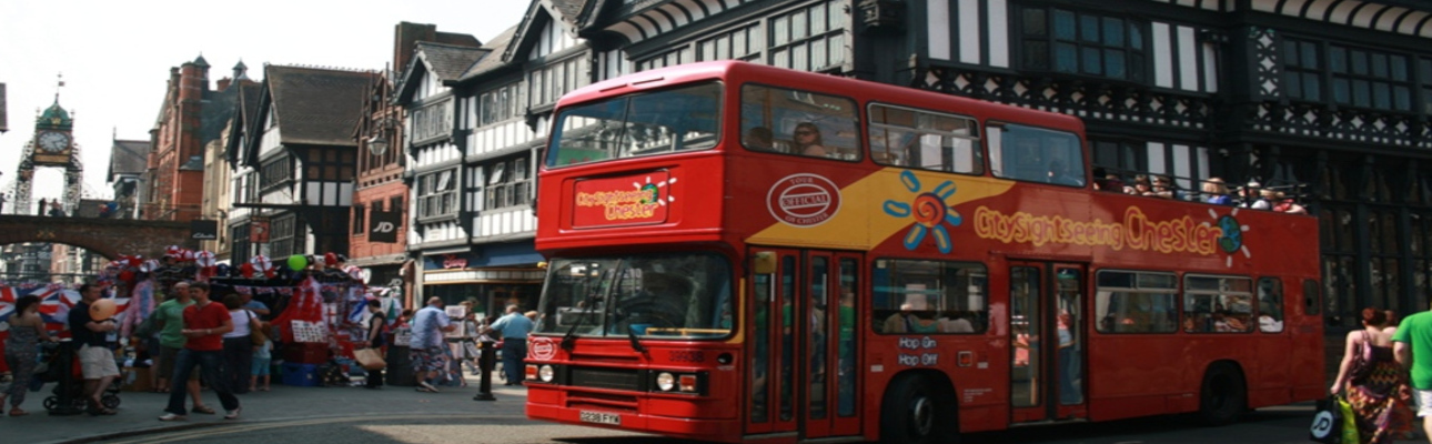 A red tour bus driving around a busy Chester street.