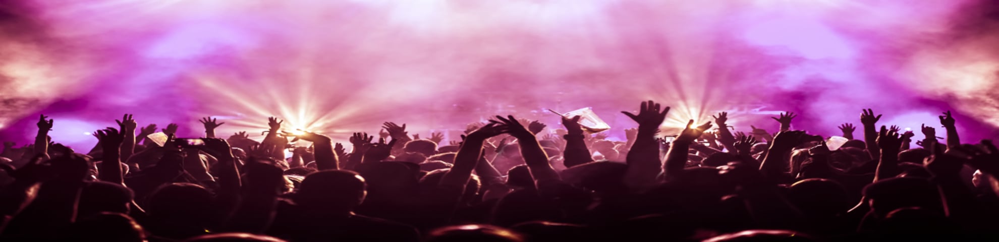 Crowd enjoying a concert with pink lighting and smoke, with their hands in the air 