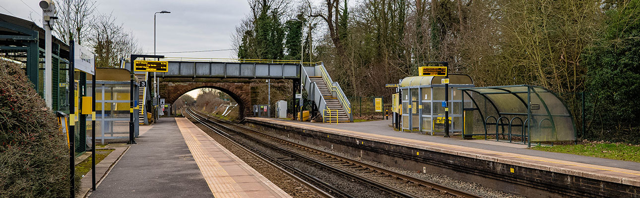 An empty platform at Capenhurst Station. There is a bridge above the train track and sheltered seating for passengers. 