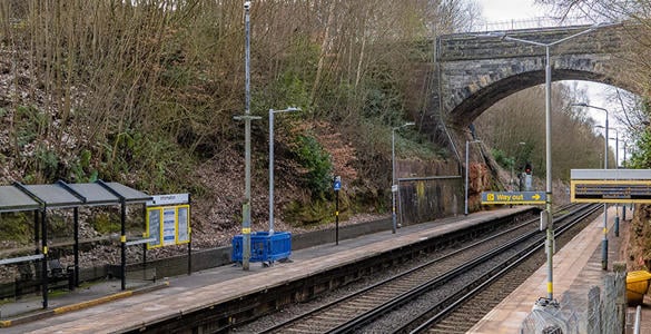 The platform at Aughton Park station. A tall tunnel bridge sits above the railway track. There is digital signage and sheltered seating on the platform. 