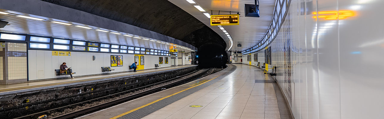 The underground station platform at Hamilton Square. Digital signage boards and lights appear. Passengers are seated waiting for a train. 