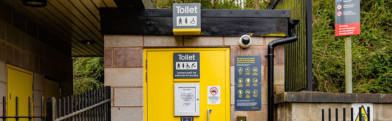 A small outdoor toilet with a yellow door and signage. There are railings nearby. 