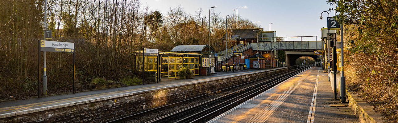 The platform at Fazakerley station. Sheltered seating, station signage appear. The station ticket office and a bridge are in the distance. 