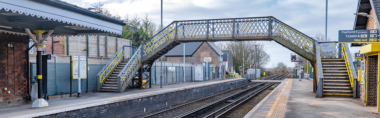 The platform at Maghull station. There are stairs and a pedestrian footbridge above the platform and railway tracks. 