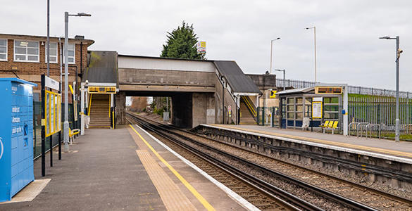 The platform and railway track at Moreton station. Sheltered seating, signage and Amazon lockers appear. The station ticket office and a bridge appear in the distance.