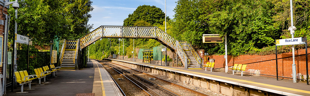 The platform at Rice Lane station. There is seating and signage on the platform. A pedestrian footbridge appears in the distance. 