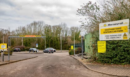 A car park at Eastham Rake station with five cars. There are trees and signage surrounding the car park. 