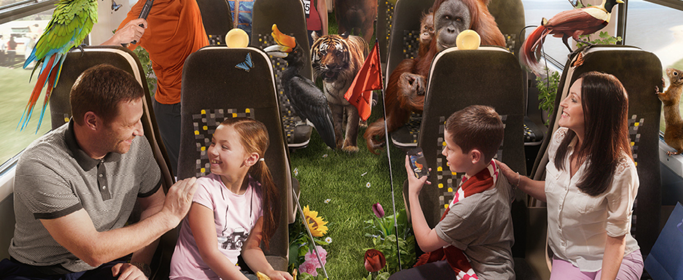 A family smiling on a train journey. There are zoo animals travelling on board behind them. 