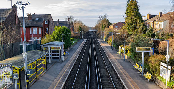 The railway tracks at Orrel Park station. There is sheltered seating and digital signage on the platform. Neighbouring houses appear in the distance. 