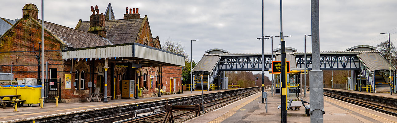 The platform at Hooton station. A pedestrian footbridge and sheltered seating appear. 