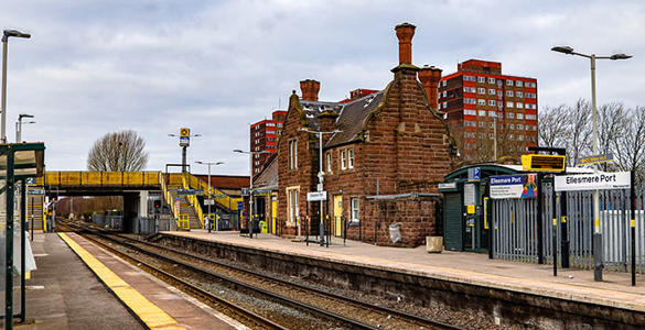 The platform and railway tracks at Ellesmere Port station. The station building, a footbridge and sheltered seating appear. 