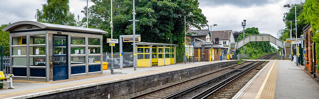 The platform at Freshfield station. Sheltered seating and signage appears. A pedestrian footbridge is in the distance. 