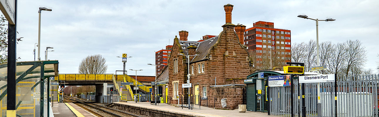 The platform and railway tracks at Ellesmere Port station. The station building, a footbridge and sheltered seating appear. 
