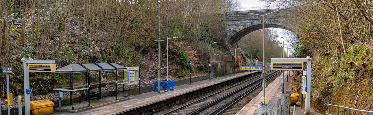 The platform at Aughton Park station. A tall tunnel bridge sits above the railway track. There is digital signage and sheltered seating on the platform. 