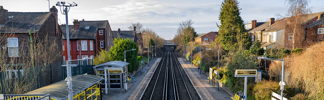 The railway tracks at Orrel Park station. There is sheltered seating and digital signage on the platform. Neighbouring houses appear in the distance. 