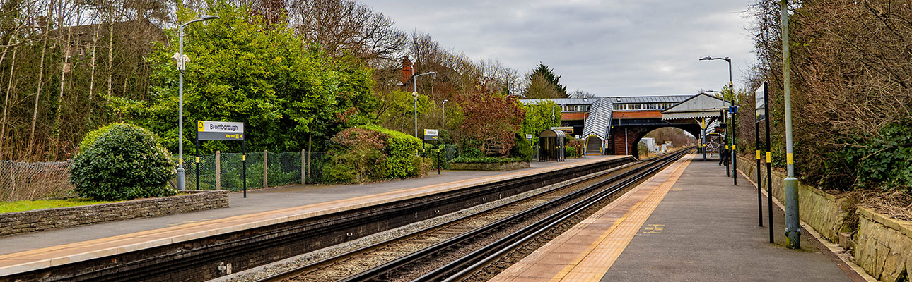 An empty platform at Bromborough station surrounded by bushes and trees. There are lampposts and a stairway to the ticket office. 