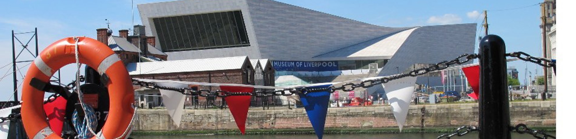 The exterior of the museum at daytime. There are flags and a lifebuoy on a railing infront of the building.